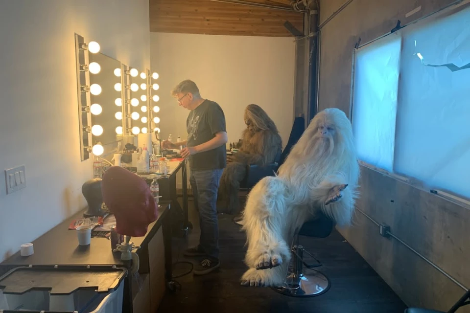 Fur-covered creature in makeup chair, attended by a man, with another creature in the background, inside a brightly-lit indoor studio.